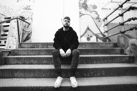 Junger Mann mit Bart und kurzen Haaren sitzt auf einer Treppe, hinter ihm eine mit Graffiti besprayte Wand und Tür, schwarzweiß